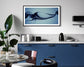 Modern kitchen with blue cabinets, a sleek marble countertop, and black appliances. A large plant stands by a round table with bowls, beneath abstract art.