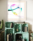 abstract heart in pink, green, gold, teal, red on white background in tan frame with tan matte on light gray wall above light teal colored crates with books and two bottles