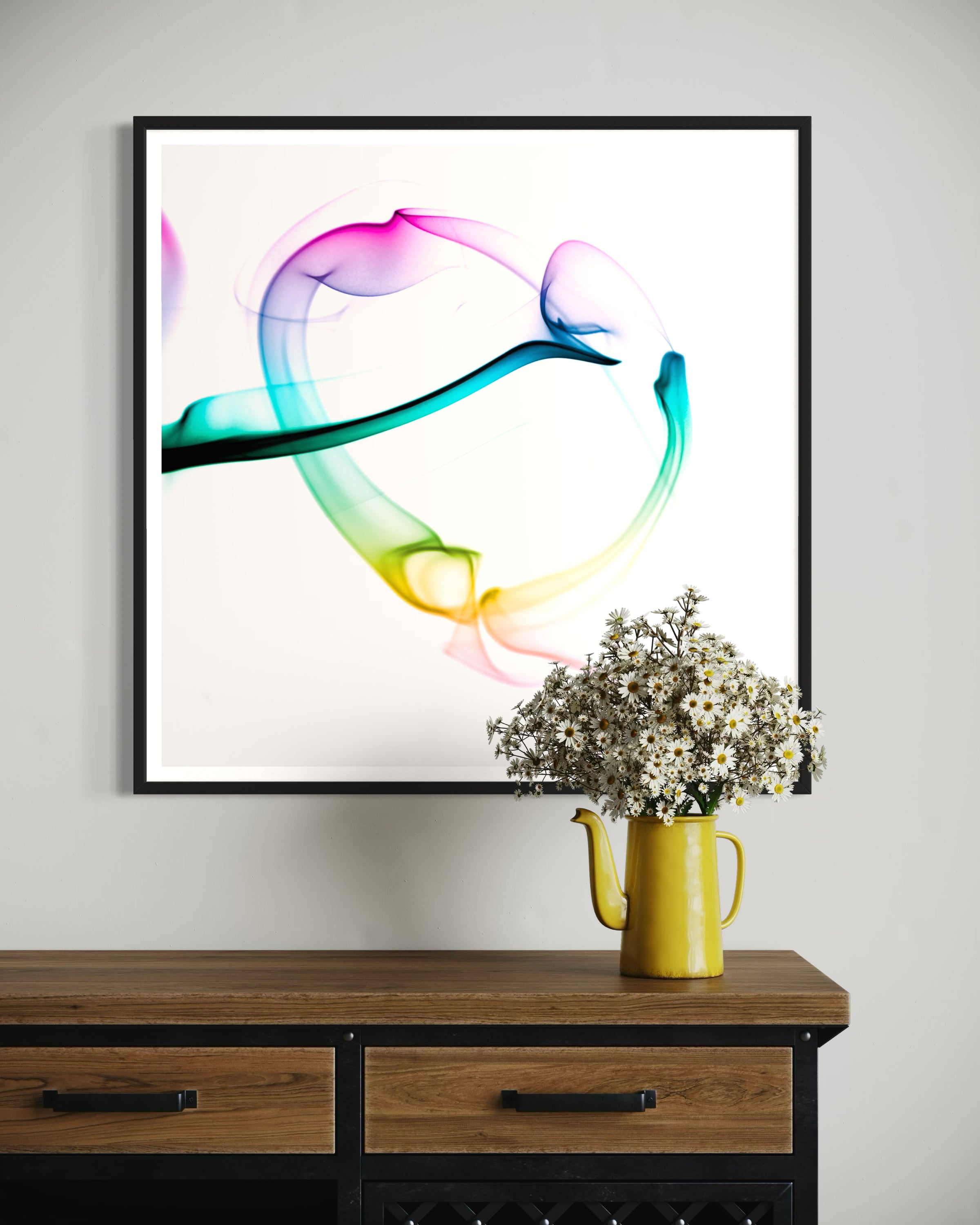 abstract heart in pink, green, gold, teal, red on white background in black frame on light gray wall above wooden table with drawers