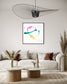 abstract heart in pink, green, gold, teal, red on white background in black frame with white matte on light gray wall above light tan sofa in earth toned living room
