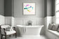 abstract heart in pink, green, gold, teal, red on white background in dark gray frame with light gray matte on light gray wall above free standing bathtub in bathroom with white and gray tones