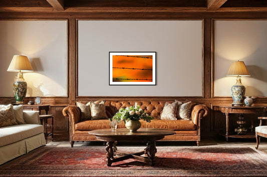 Close up of barbed wire fence with sunset in the background, in black frame with white matte on tan wall above brown leather couch in rustic living room