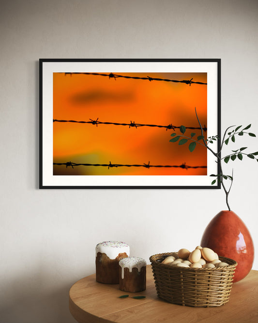 Close up of barbed wire fence with sunset in the background, in black frame with white matte on tan wall above small table with two small cakes, large basket of eggs and orange vase with twig in it