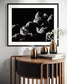 black and white photo of clouds, one looks like a dog's head in profile in black frame with white matte on white wall above small wooden table
