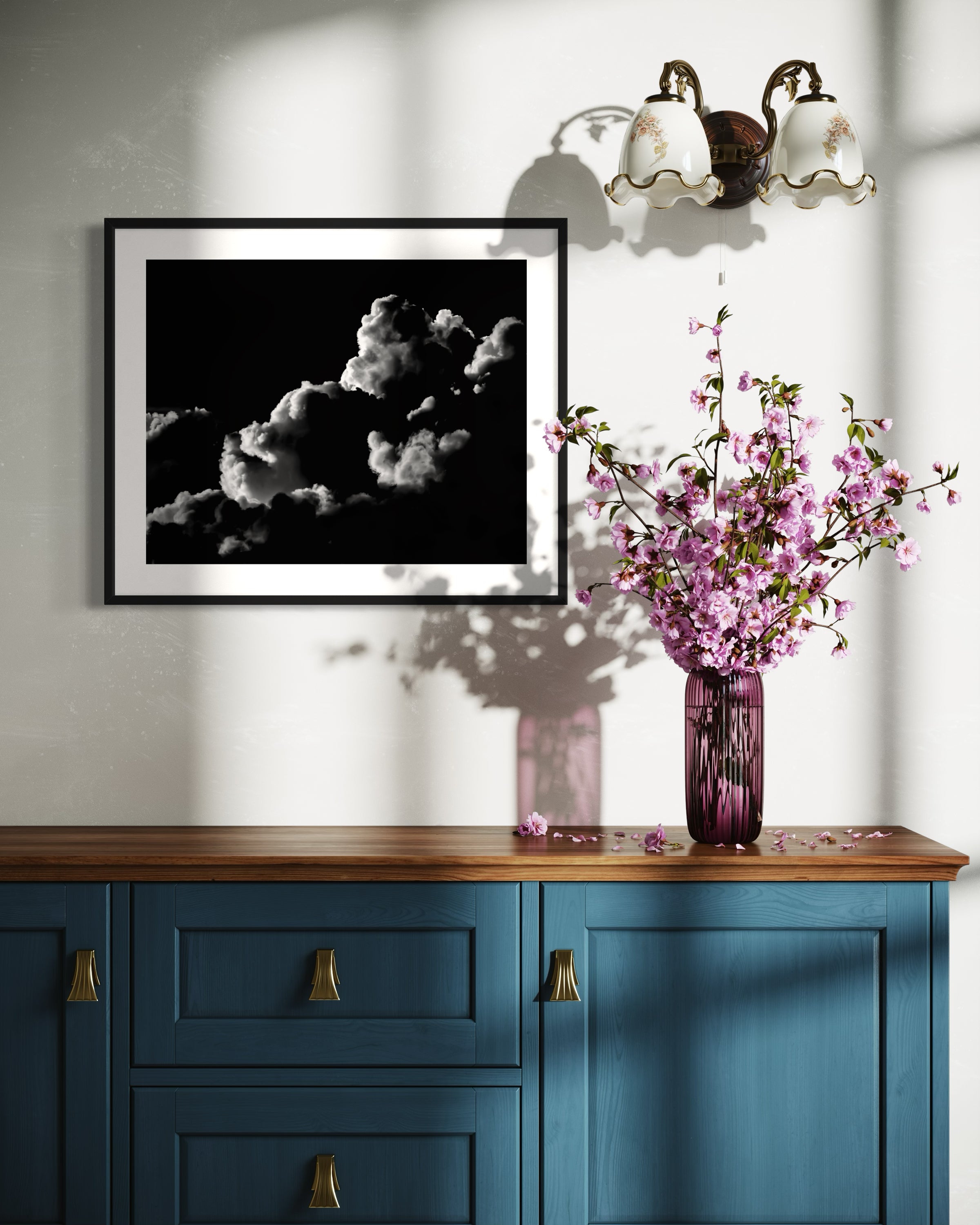 black and white photo of clouds, one looks like a dog's head in profile in black frame with white matte on white wall above blue piece of furniture with flowers on top