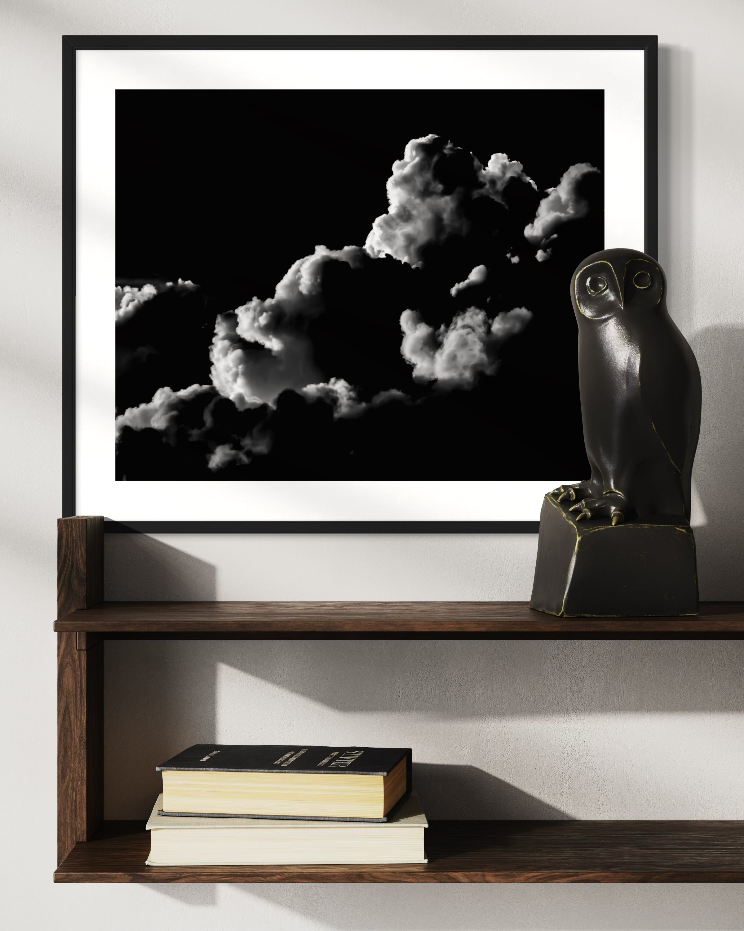 black and white photo of clouds, one looks like a dog's head in profile in black frame with white matte on white wall above wooden shelf with owl sculpture on top