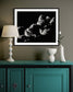 black and white photo of clouds, one looks like a dog's head in profile in black frame with white matte on warm gray wall above teal piece of furniture with lamp on top