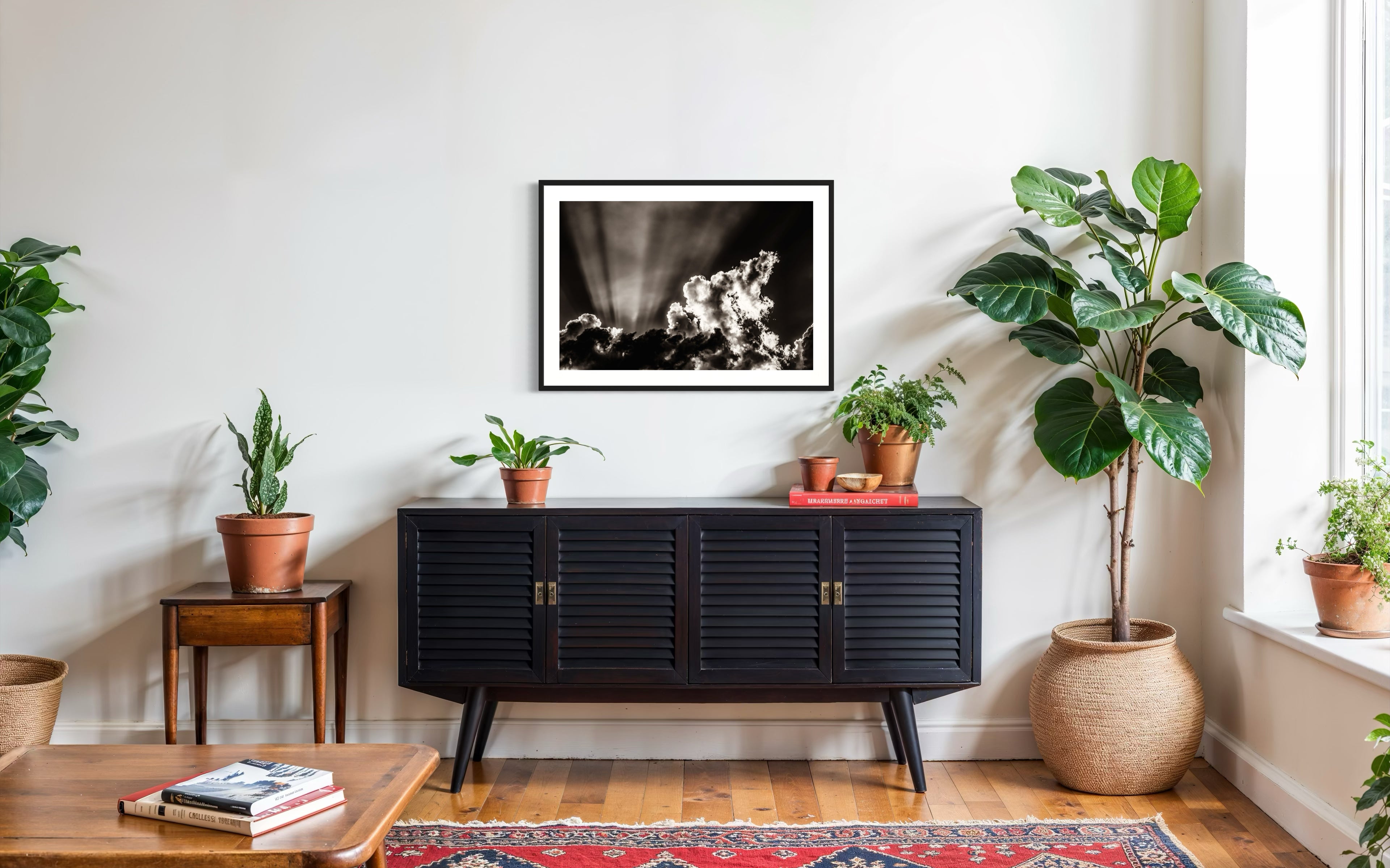 black and white photo of dramatic sky with some clouds and rays of light  in black frame with white matte on white above piece of black furniture with tall green plants on the side
