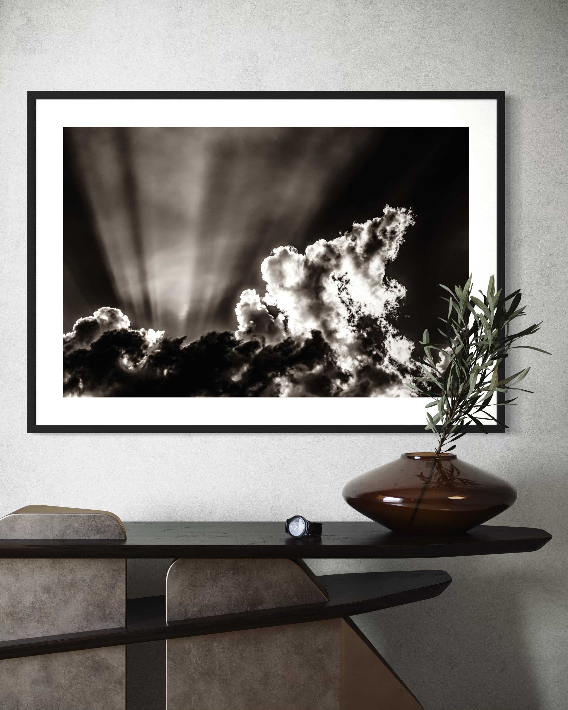 black and white photo of dramatic sky with some clouds and rays of light  in black frame with white matte on light gray wall above small funky table with single plant branch in wide brown vase