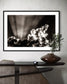 black and white photo of dramatic sky with some clouds and rays of light  in black frame with white matte on light gray wall above small funky table with single plant branch in wide brown vase