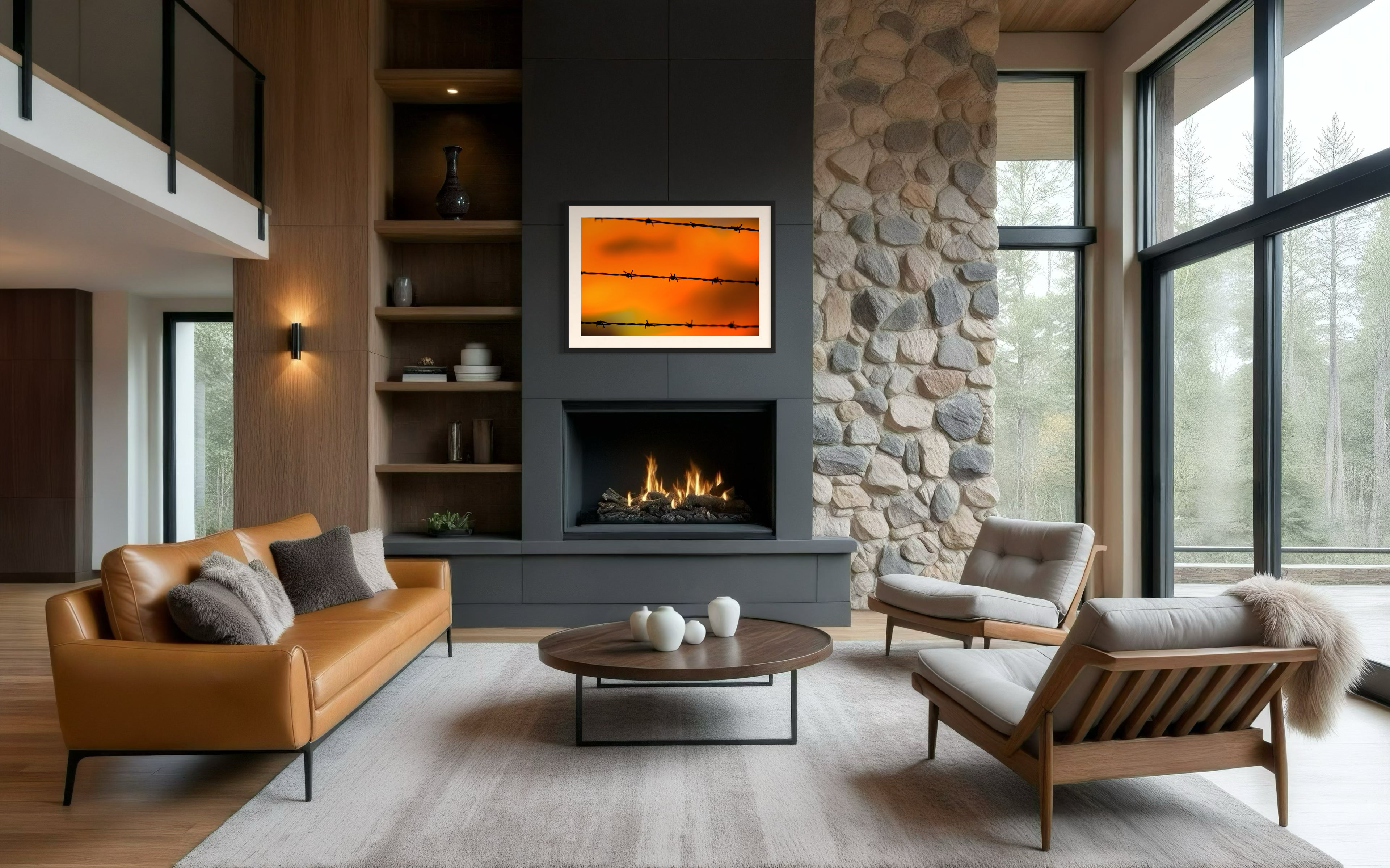 Close up of barbed wire fence with sunset in the background, in black frame with white matte on dark gray wall above fireplace in rustic living room with fireplace and floor to ceiling windows on the right