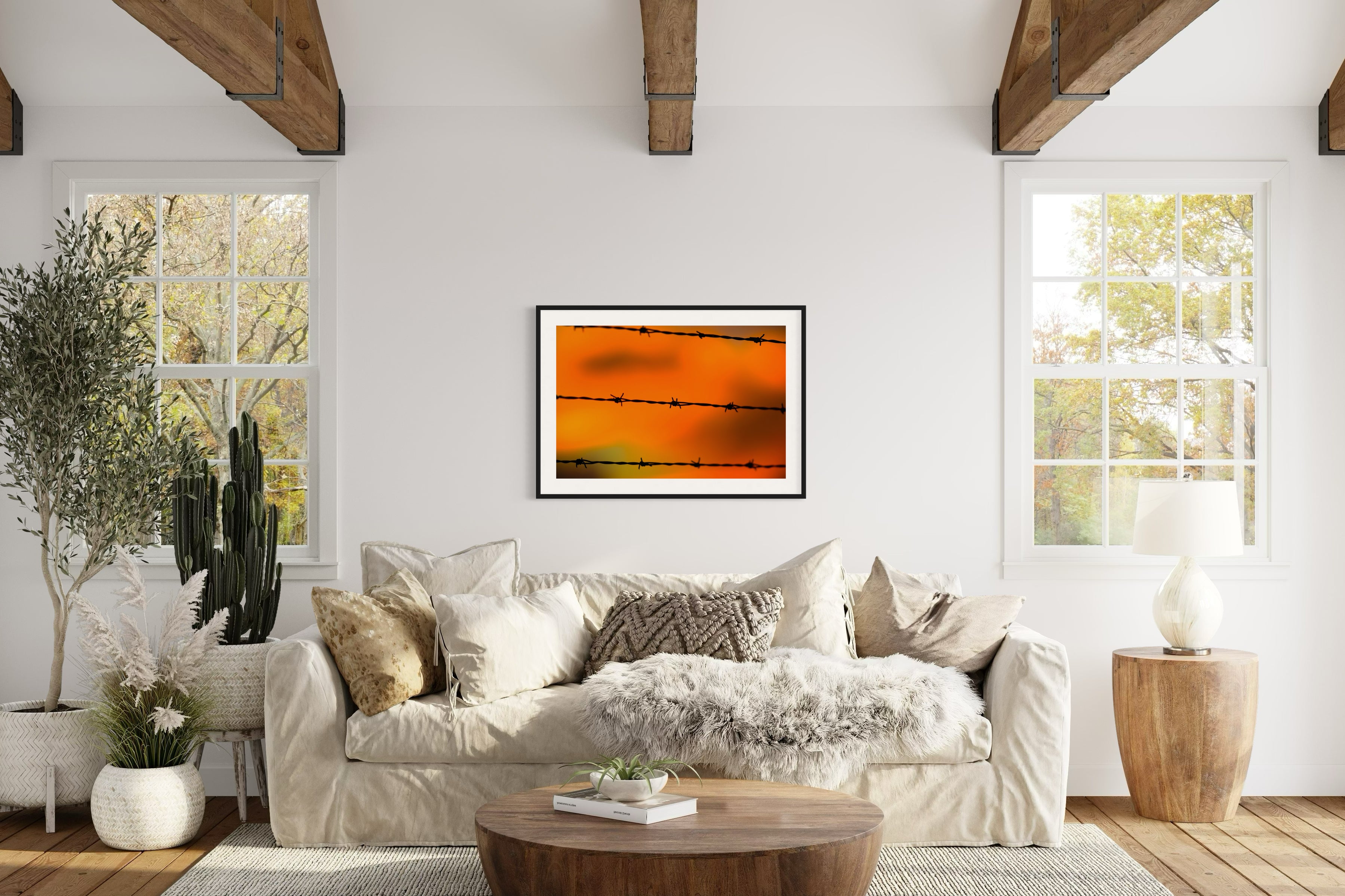 Close up of barbed wire fence with sunset in the background, in black frame with white matte on off white wall above couch on tan wall in rustic living room with beams in the ceiling and hardwood floors