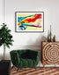 swirls of blue, yellow, orange, teal, purple and red on light yellow background in black frame with off white matte on light tan wall in modern sitting room with dark green chair and tall green plant
