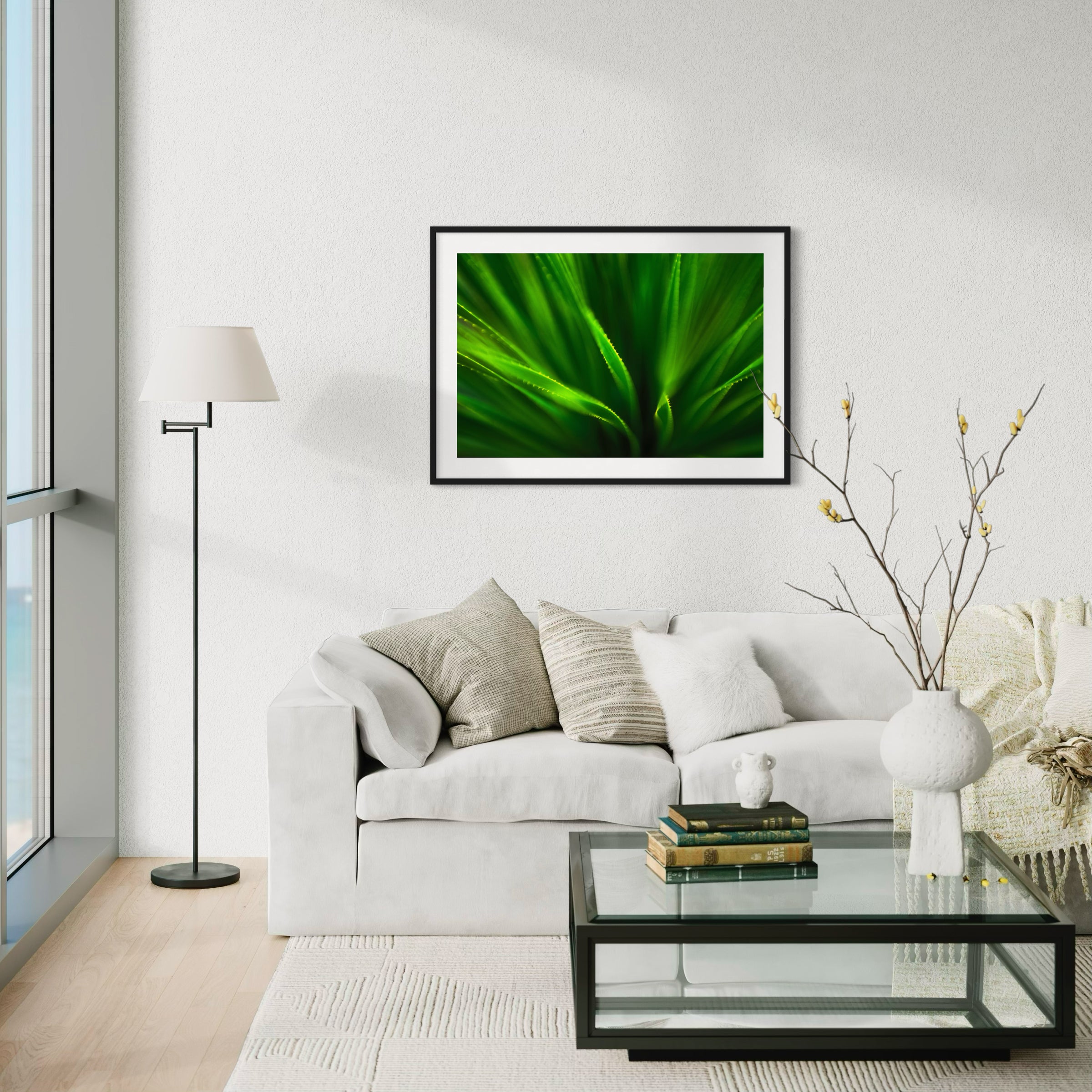 green agave like plant with selective focus in black frame with white matte on off white wall above white couch in modern living room with mostly white tones