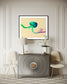 photo of abstract purple, green, blue, yellow wave at the bottom on orange overall in black frame with white matte on white wall above small warm gray piece of furniture in upscale home