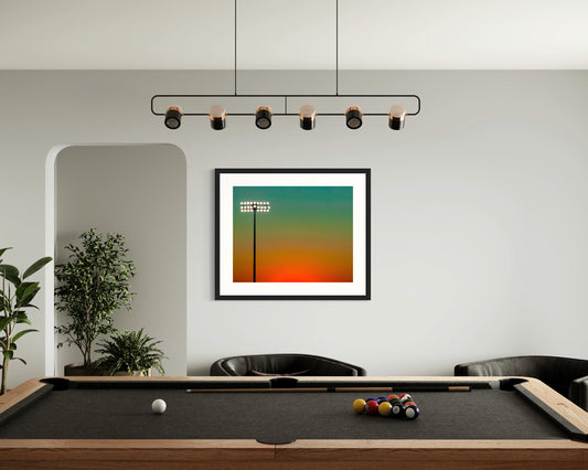  photo of sunset at football stadium single pole of lights left of center, gradient sky teal blue to pale yellow to orange gradient top to bottom in black frame on wall in game room with pool table