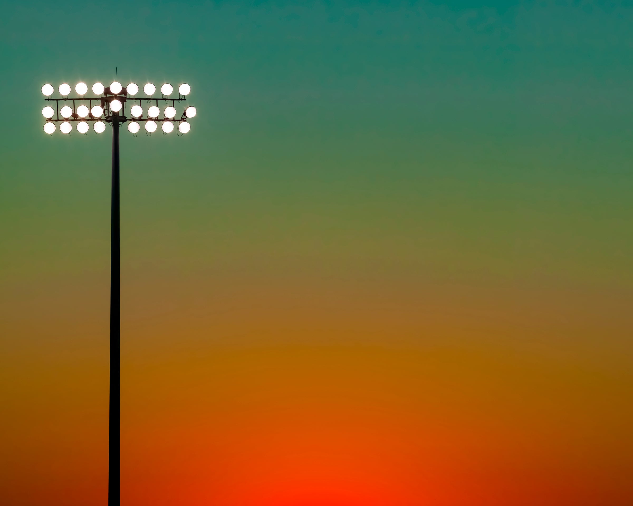  photo of sunset at football stadium single pole of lights left of center, gradient sky teal blue to pale yellow to orange gradient top to bottom