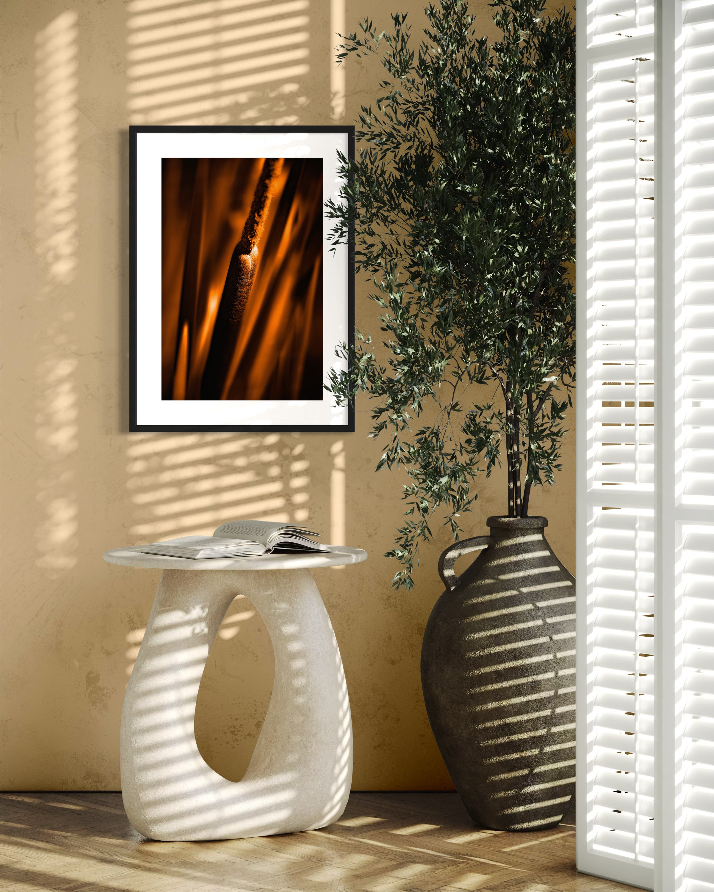 reeds in gold tone with selective focus photo in black frame with white matte on tan wall above small white table with large green plant on the right