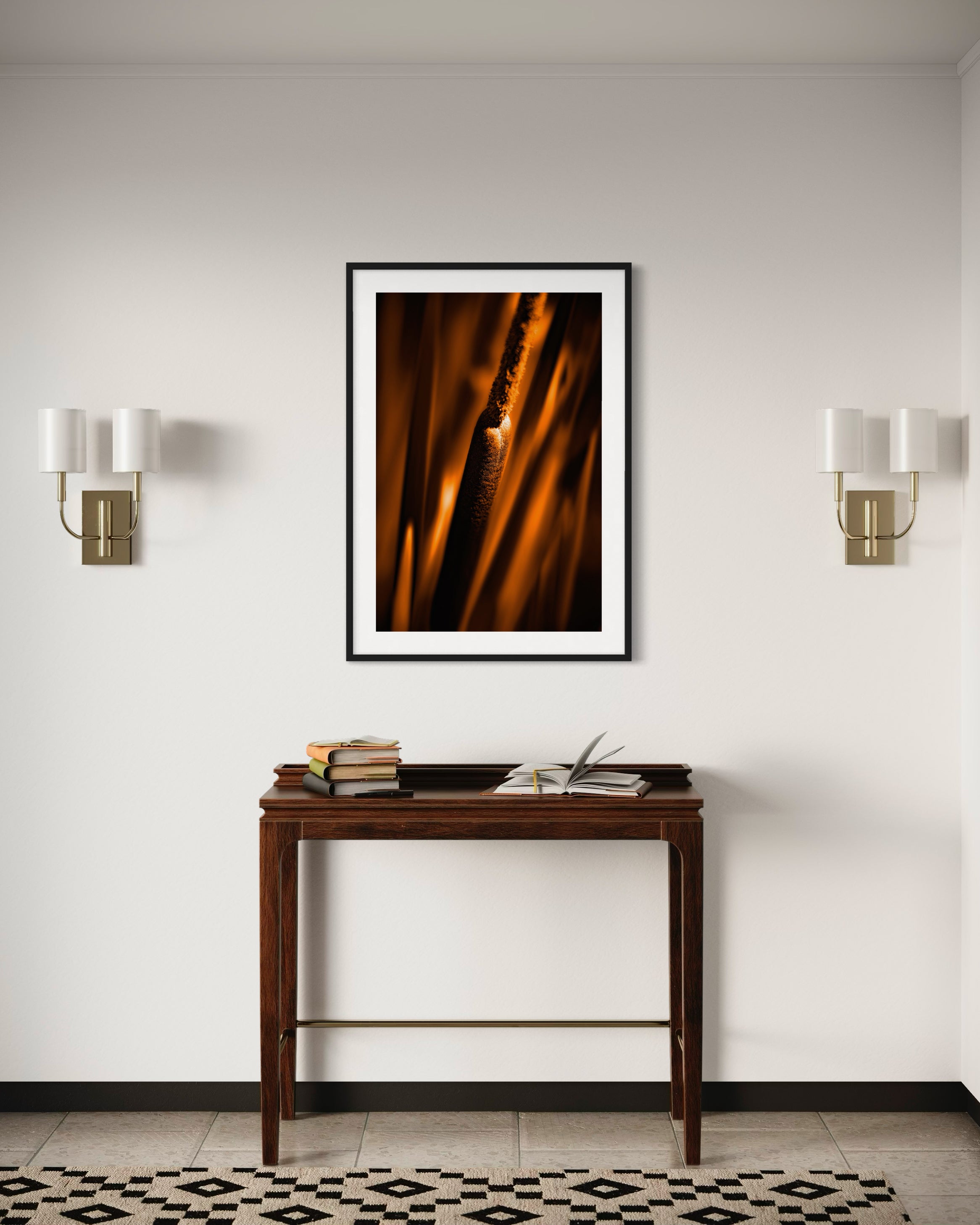 reeds in gold tone with selective focus photo in black frame with white matte on white wall above small dark wooden table, sconces on each side of photo