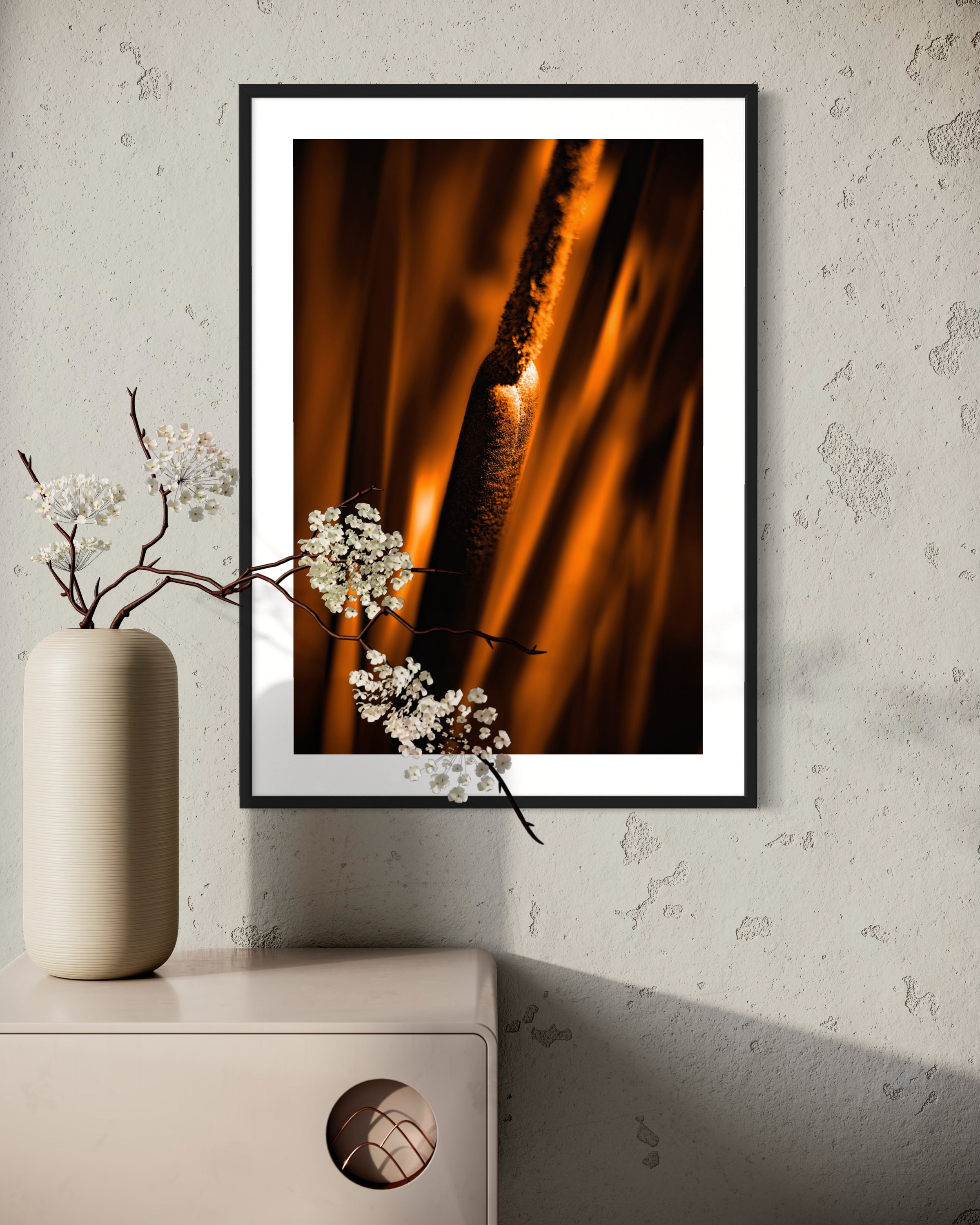 reeds in gold tone with selective focus photo in black frame with white matte on light gray concrete wall above small piece of furniture