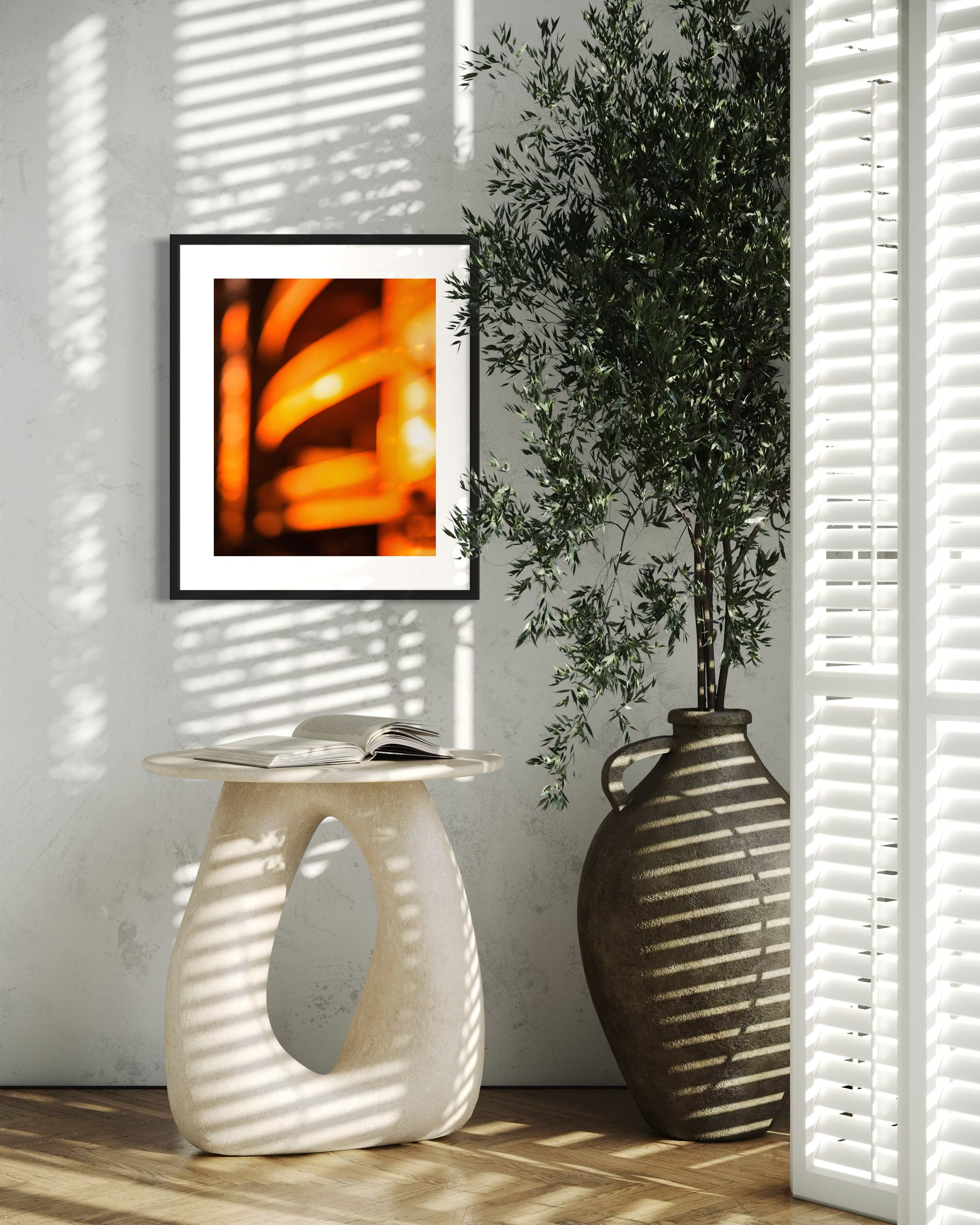 photo of abstract orange blurred lines in black frame with white matte on white wall above end table, tall green plant on the right, shadows from plant and mini blinds are on the photo