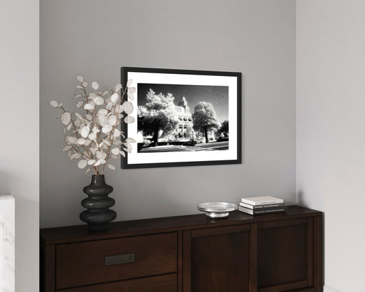 a black and white photo of a building with trees in front in black frame on gray wall above dark wooden table