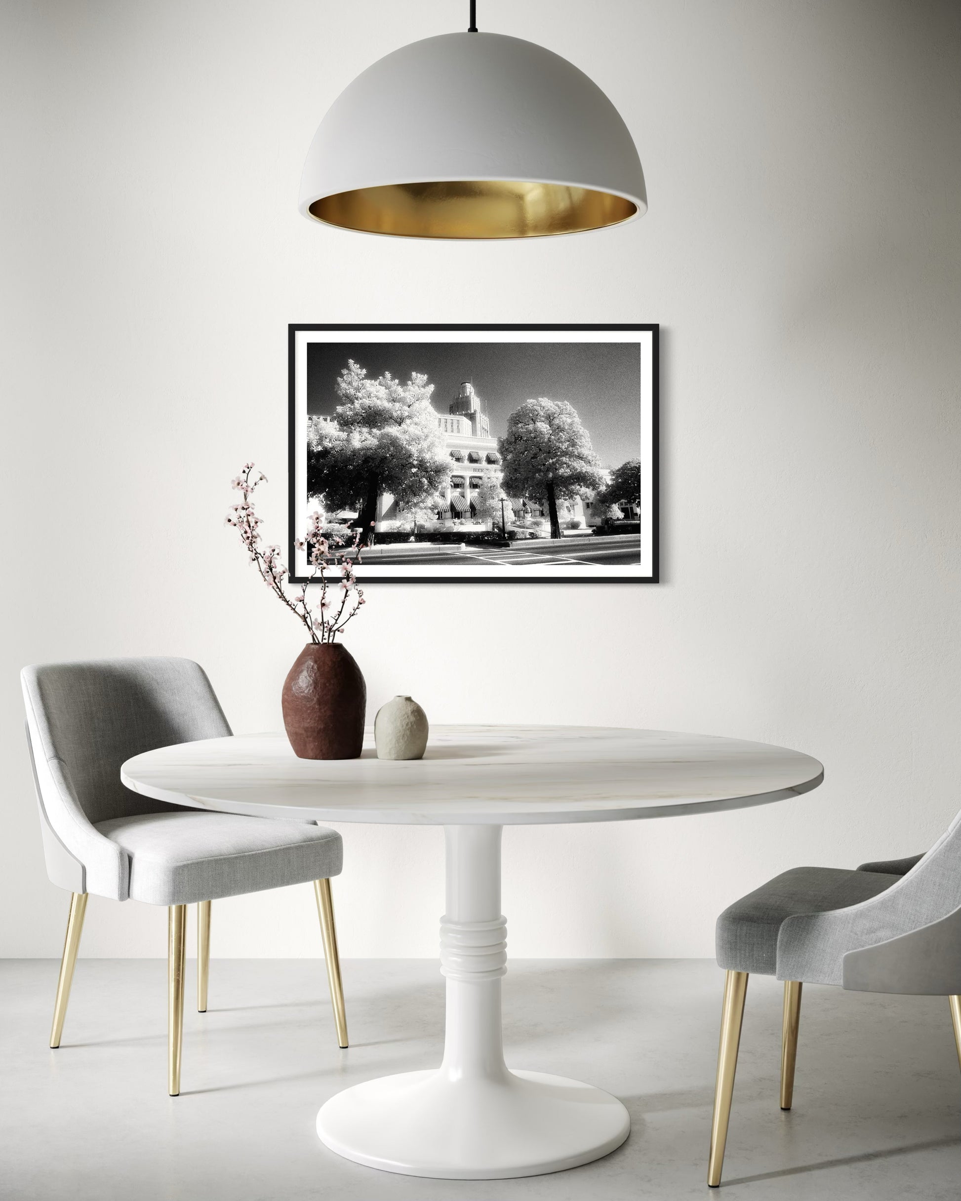 a black and white photo of a building with trees in front in black frame with white matte on white wall in minimalist breakfast room with all white except for 2 light gray chairs and a small brown ceramic vase with small pink plant branches