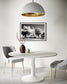 a black and white photo of a building with trees in front in black frame with white matte on white wall in minimalist breakfast room with all white except for 2 light gray chairs and a small brown ceramic vase with small pink plant branches