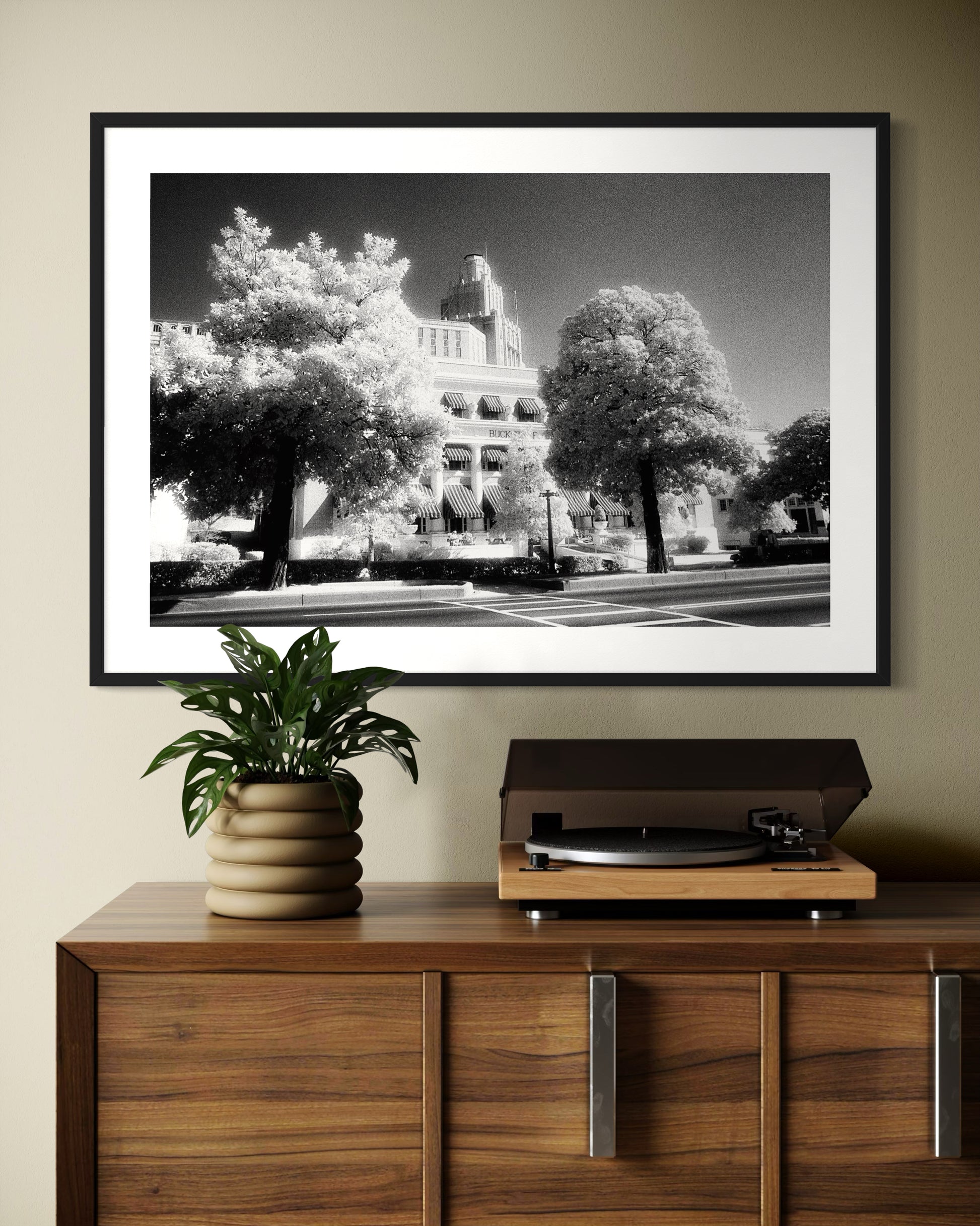 a black and white photo of a building with trees in front in black frame with white matte on tan wall above wooden table with turntable and small plant on top