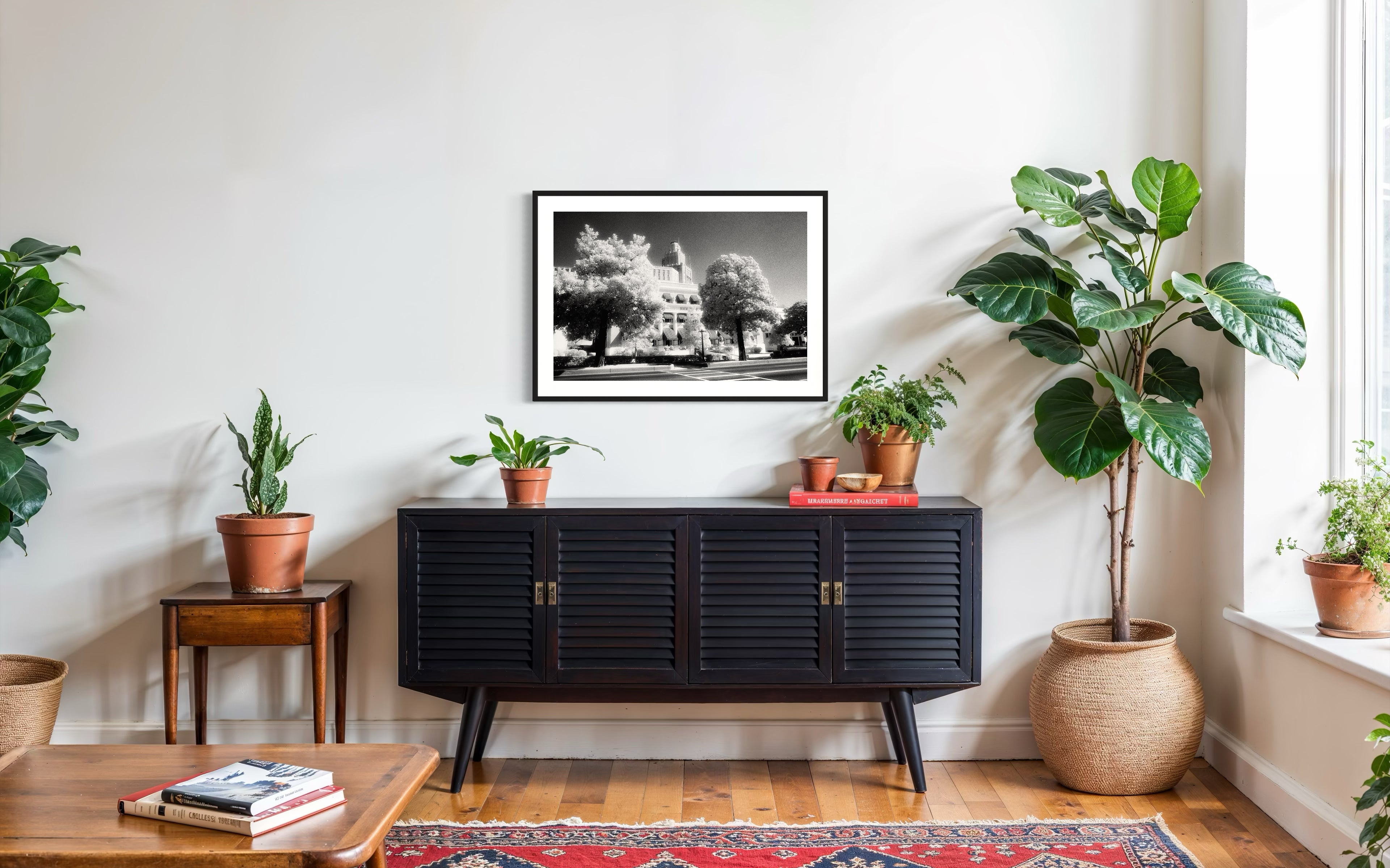 a black and white photo of a building with trees in front in black frame with white matte on white wall above black piece of furniture on hardwood floors, also a couple of tall green plants on the sides