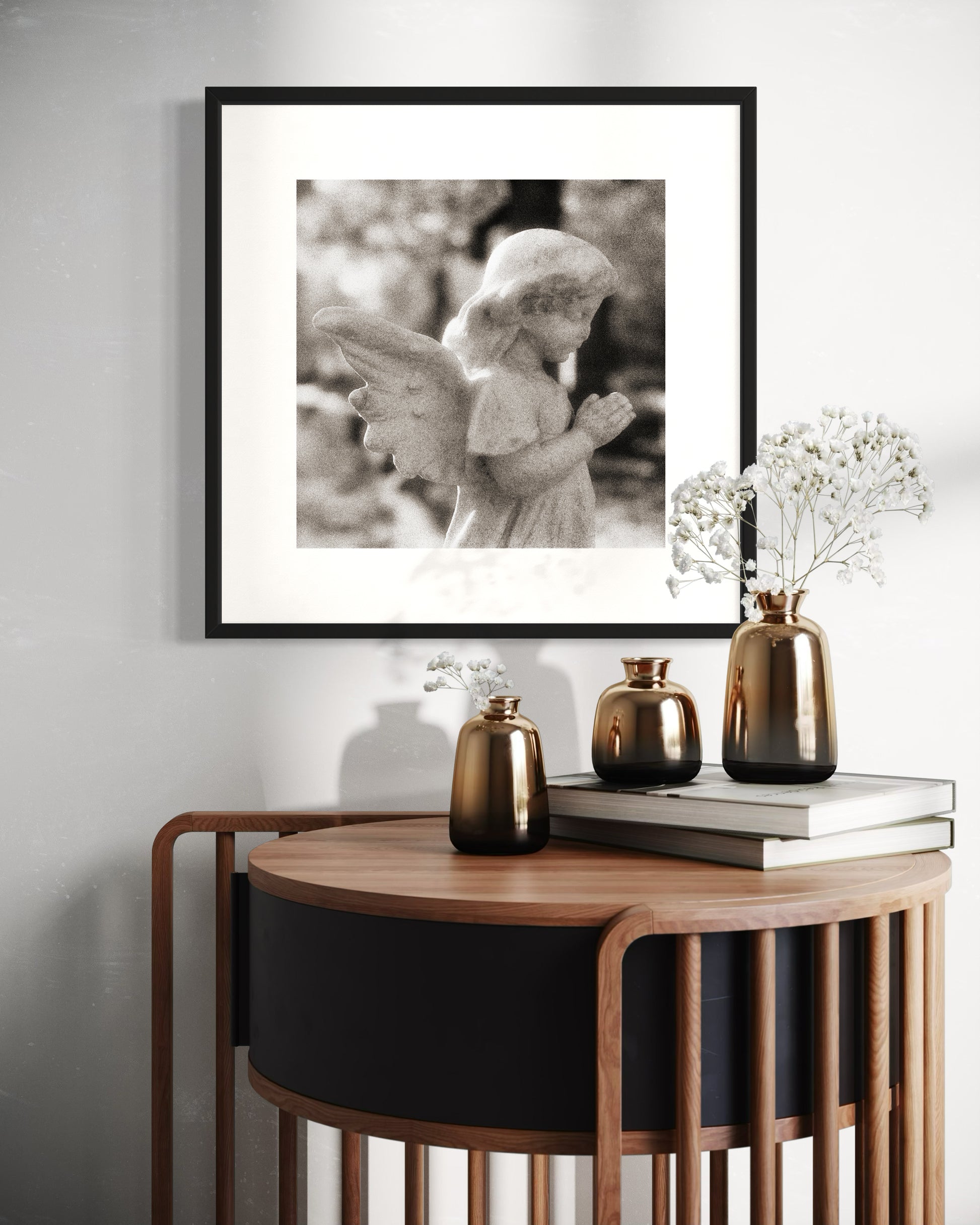 cherub statue with hands in praying position in sepia tone in black frame with white matte on white wall above modern wooden table with three vases and two books on op