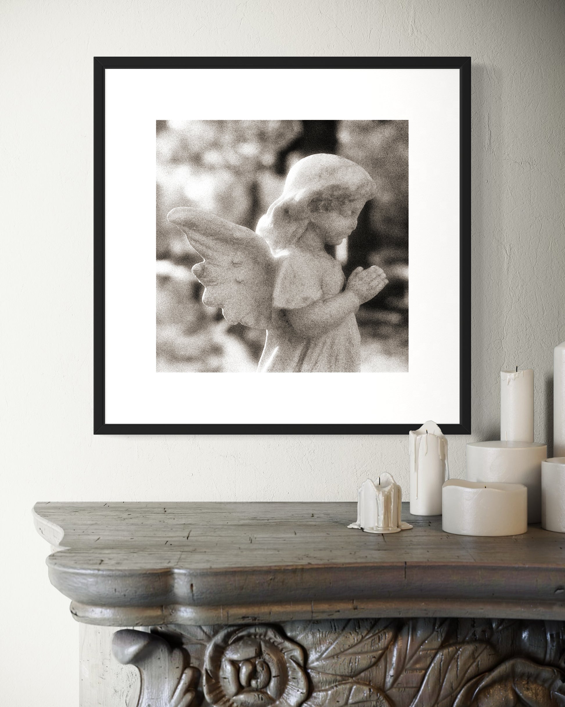 cherub statue with hands in praying position in sepia tone in black frame with white matte on light gray wall above ornate dark gray table with white candles on top