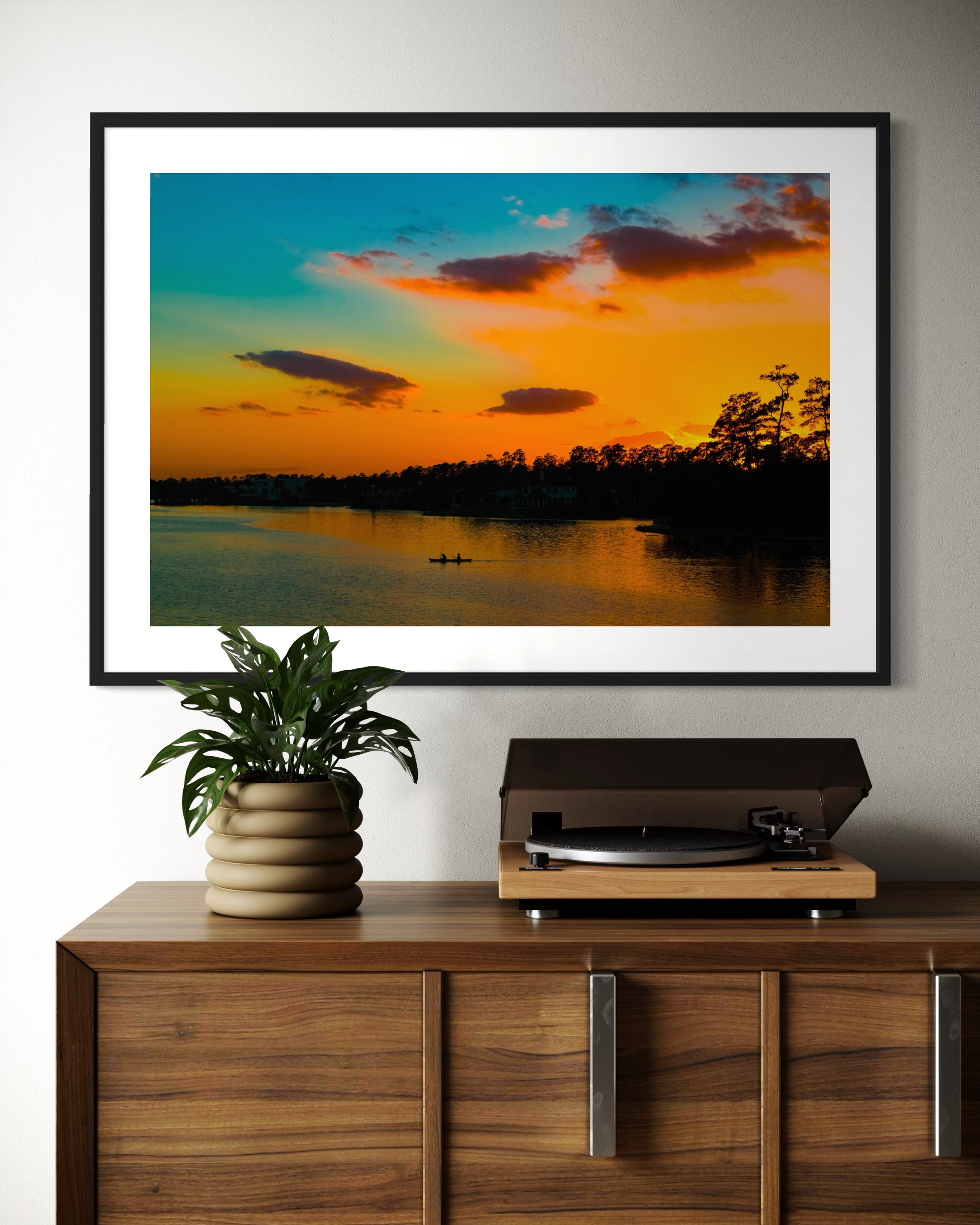 wide angle photo of kayak on lake at sunset in black frame with white matte above wooden piece of furniture with white record player on top