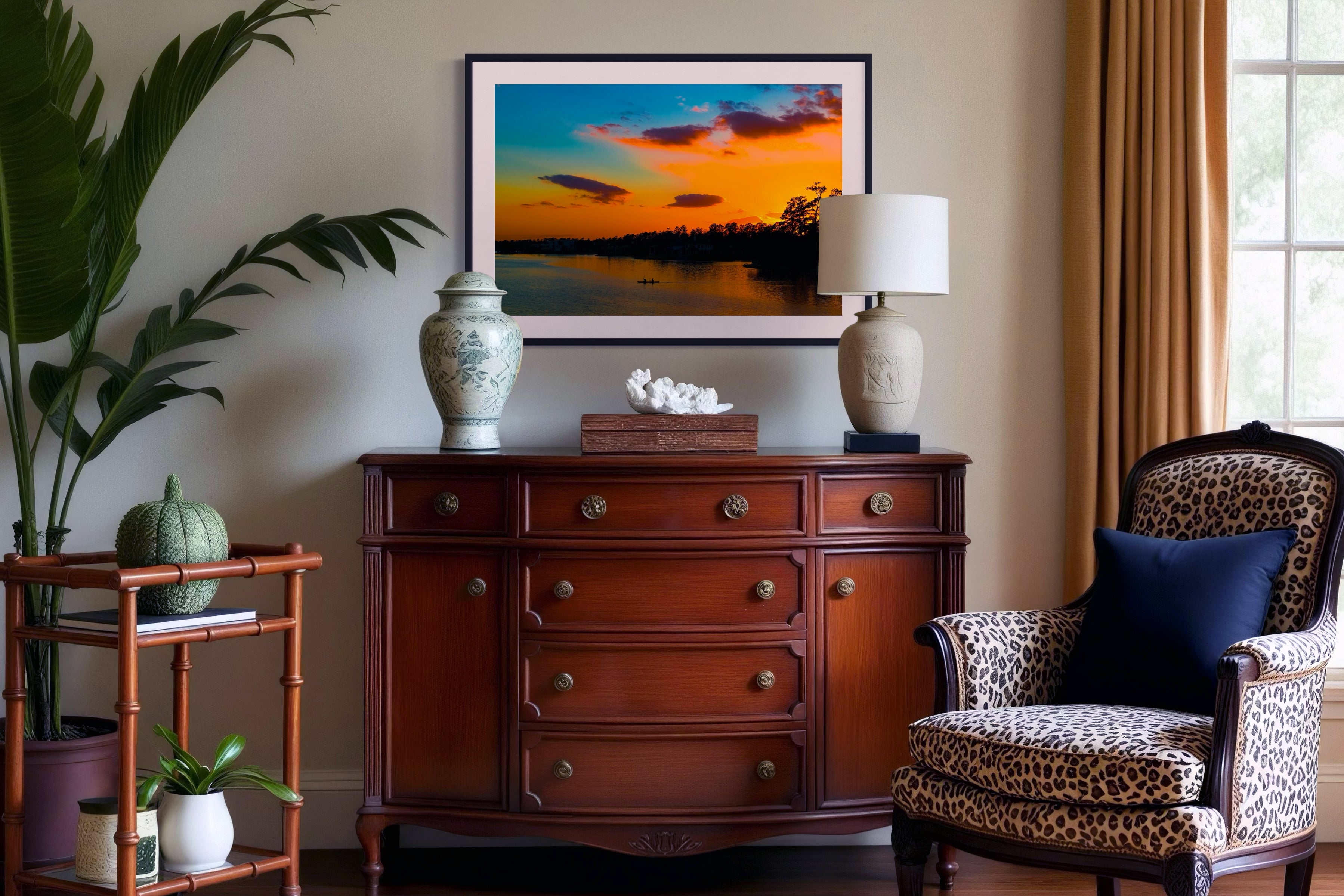 wide angle photo of kayak on lake at sunset in black frame with white matte on off white wall above wooden piece of furniture with drawers, large plant on the left side, chair with cheetah spots fabric