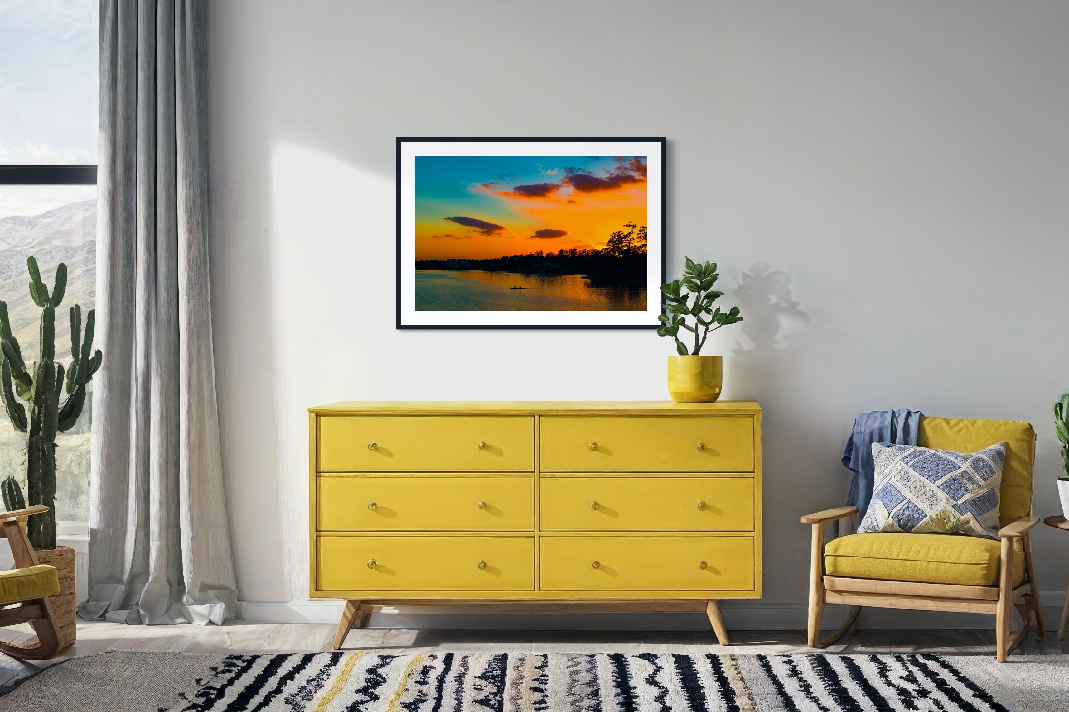wide angle photo of kayak on lake at sunset in black frame with white matte on off white wall in sitting area with yellow chair and yellow piece of furniture with drawers