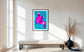 bright pop art photo of pink lily on blue background in black frame with white matte above light tan long bench with cushion  in modern sitting room with white and light tan tones