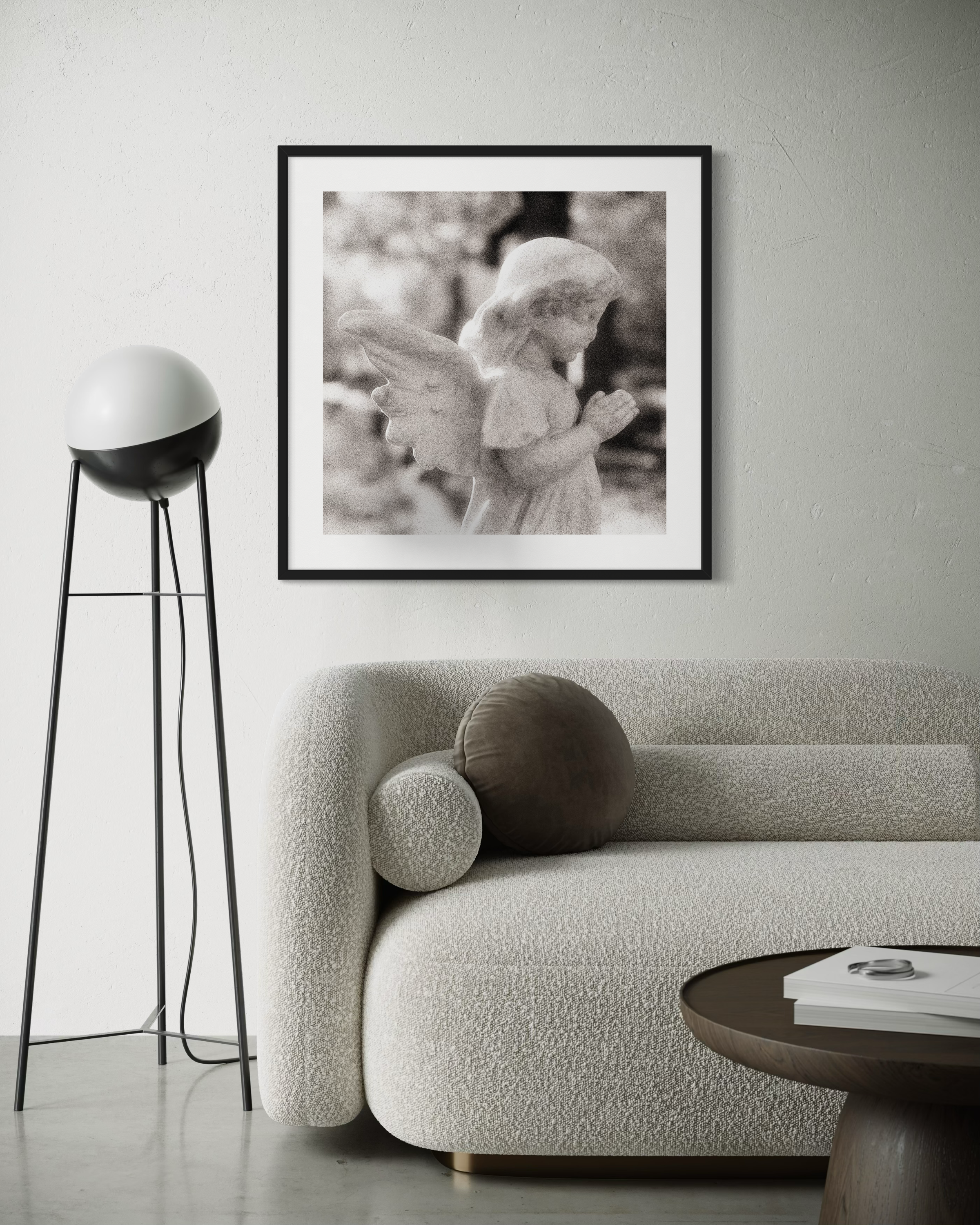 cherub statue with hands in praying position in sepia tone in black frame with white matte on light gray wall in modern room with couch and white sphere lamp with black legs on the concrete floor