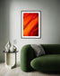 orange abstract with upward motion in black frame with white matte on light gray wall above dark green puffy modern couch with end table
