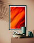 orange abstract with upward motion in black frame with white matte on tan wall with palm tree shadow on part of the print above table
