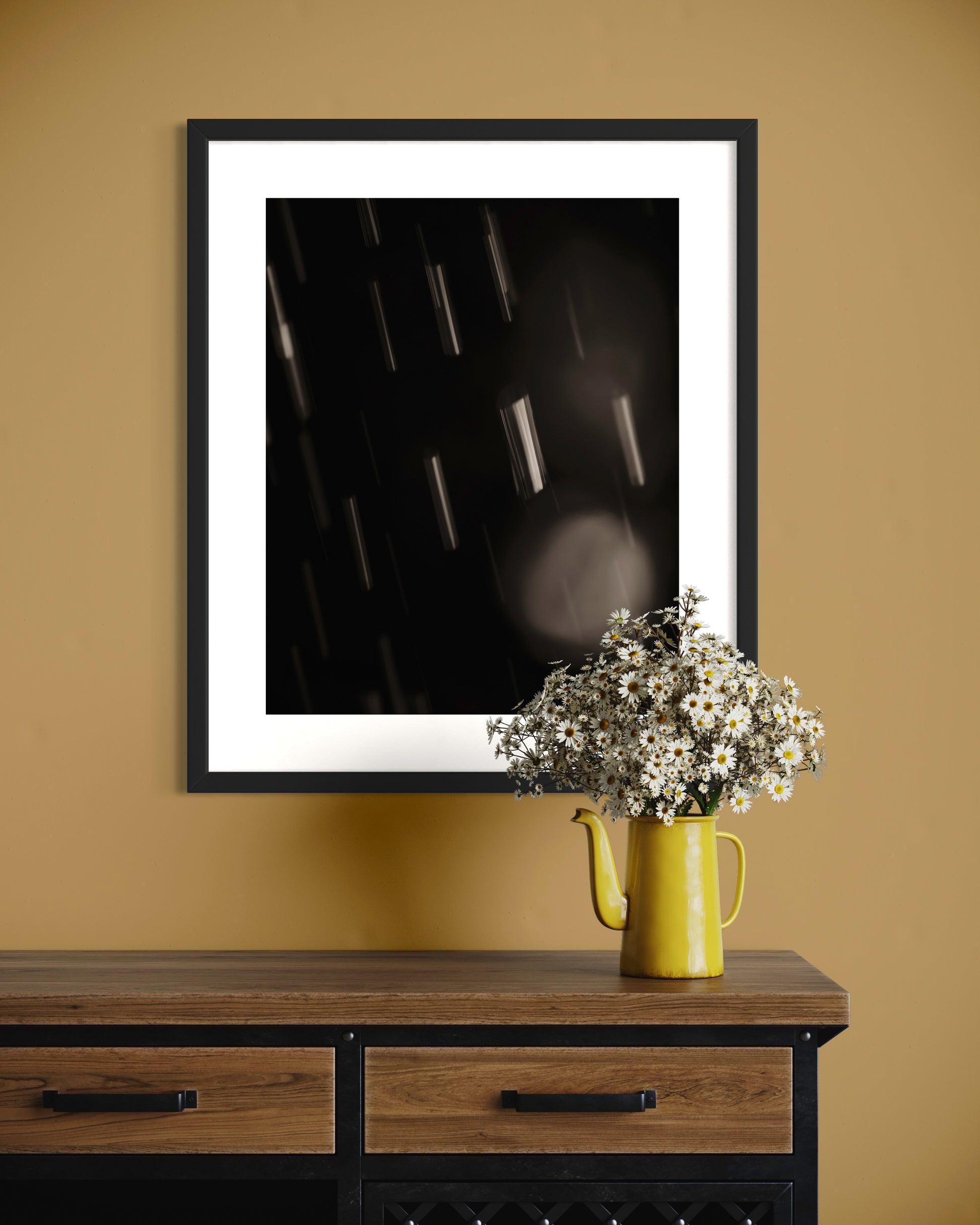 close up warm toned photo of streaks of rain in black frame on pastel yellow wall with flowers on table below