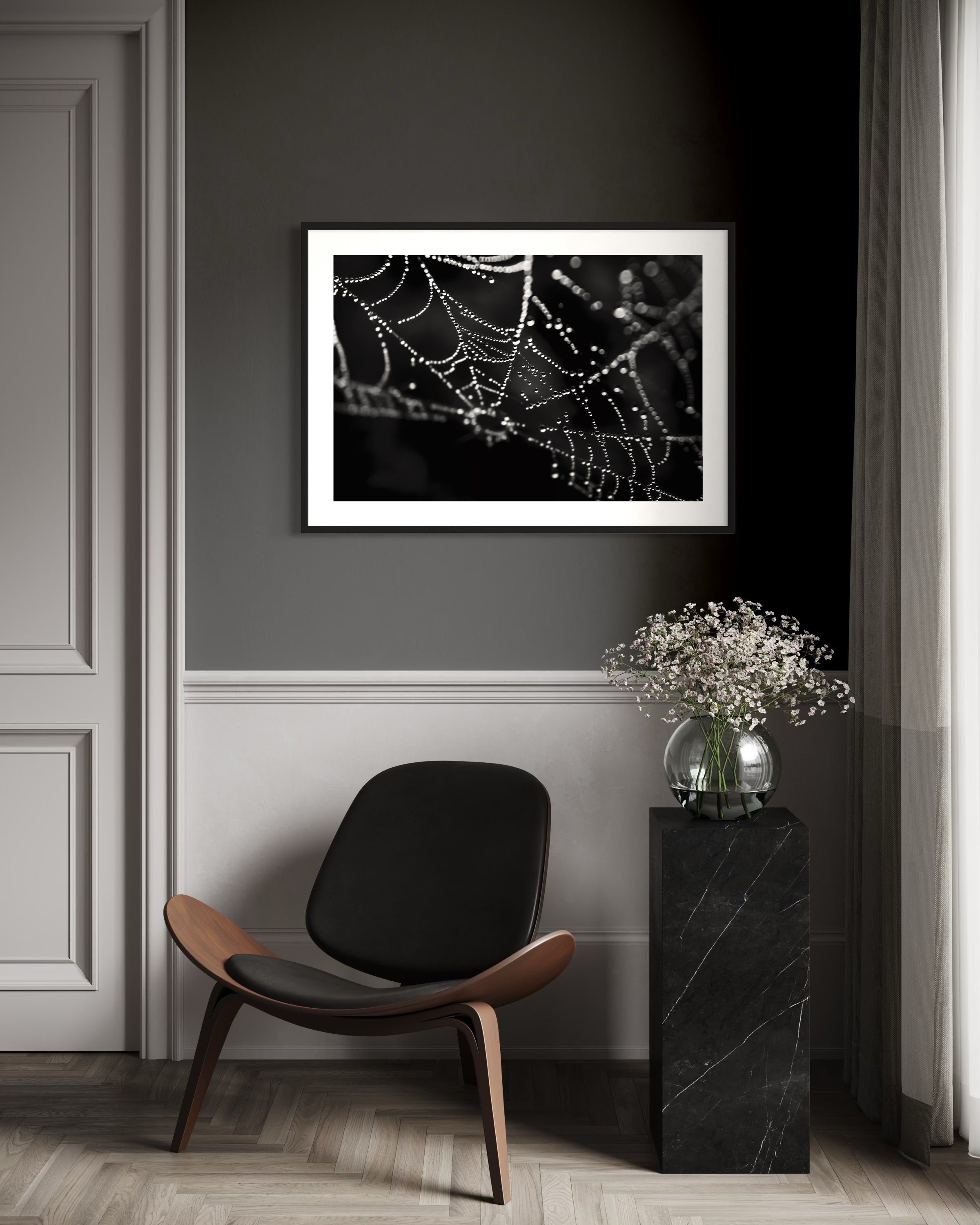 Framed black and white photograph of a spider web on a wall in a room with a chair and vase.