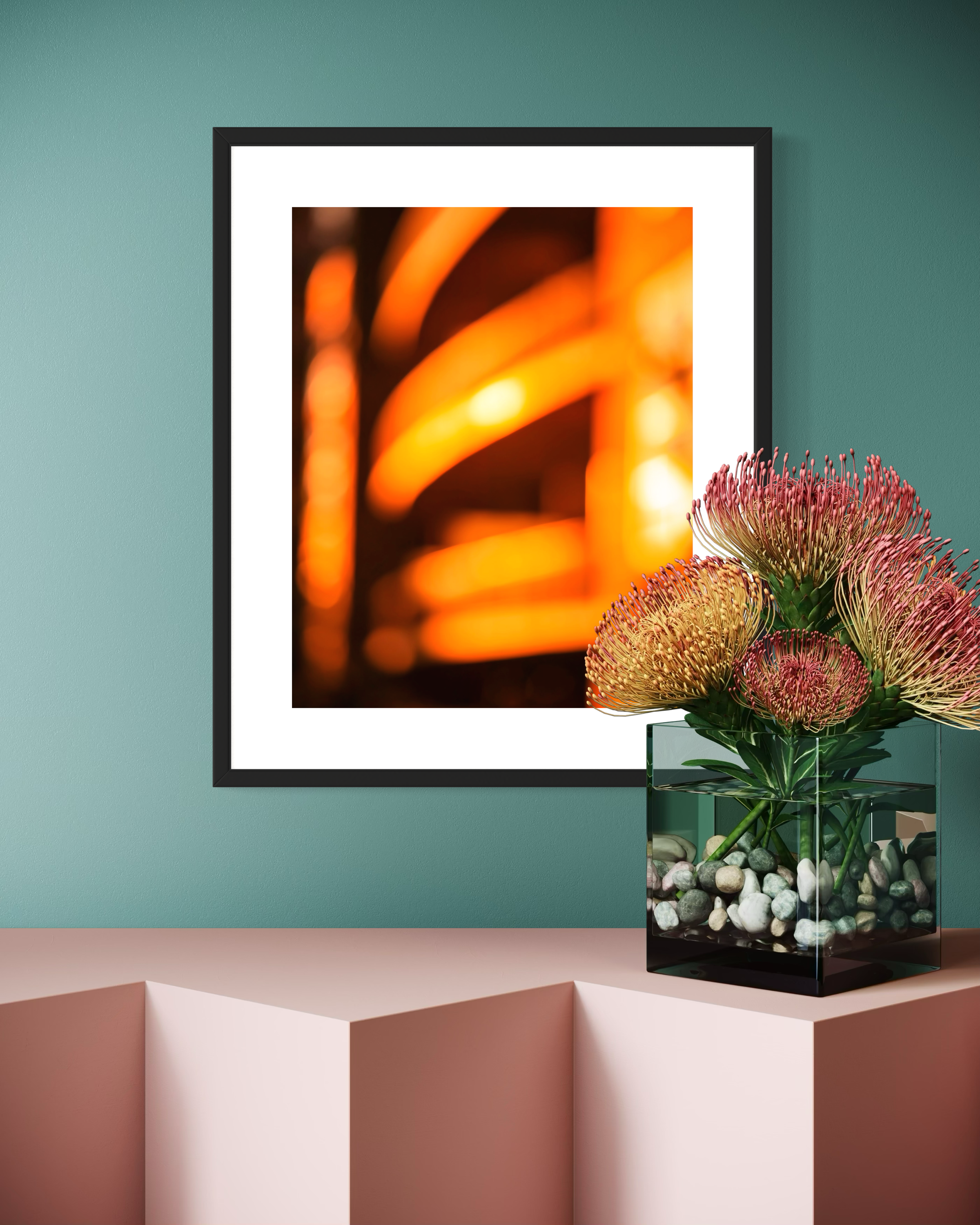 photo of abstract orange blurred lines in thick black frame with white matte on pastel green wall above pastel small angular table with flowering plant in water 