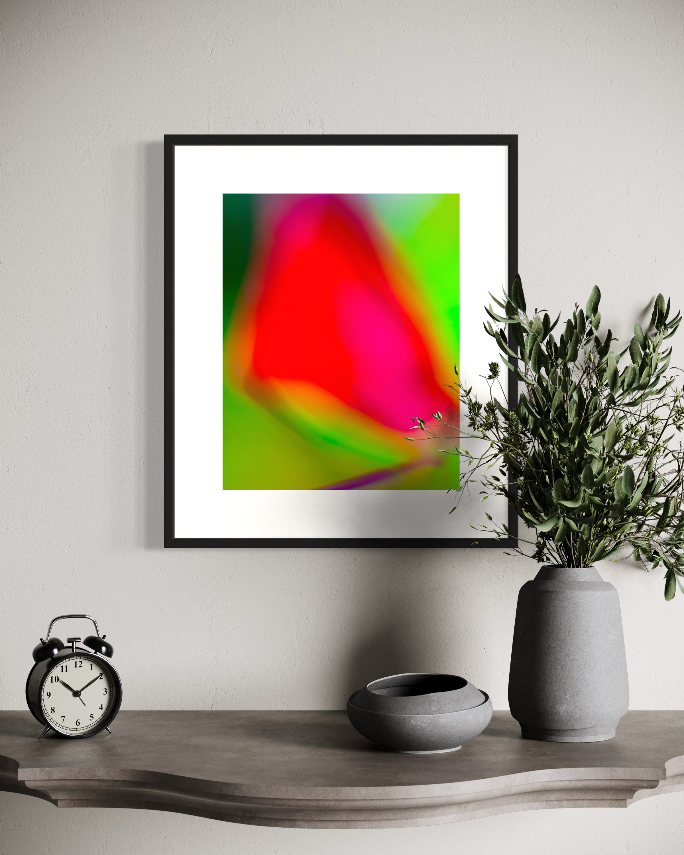 photo of blurry red leaf on blurred bright green background in black frame with white matte on white wall above small shelf with old clock and plant on top