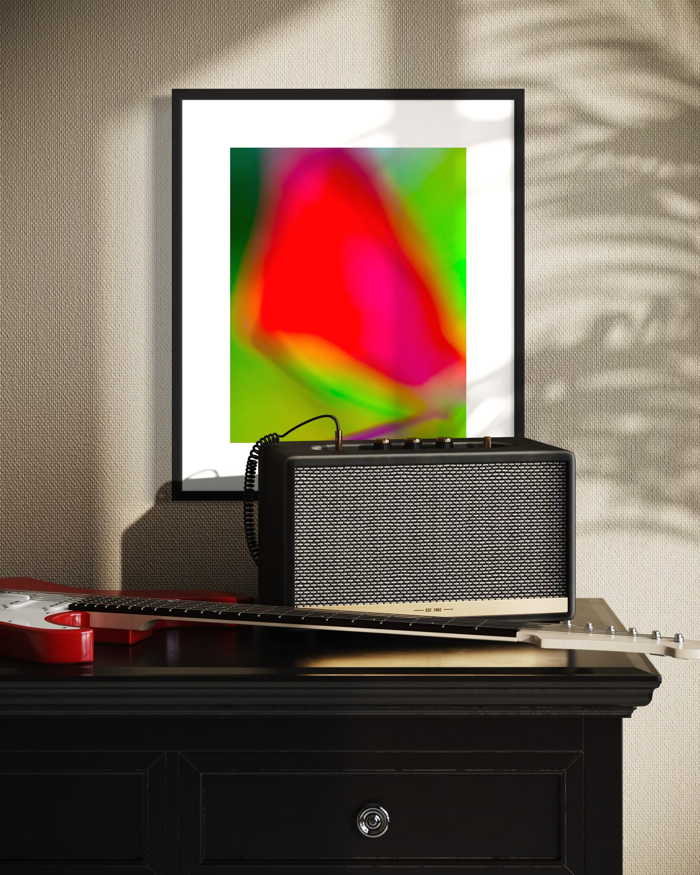 photo of blurry red leaf on blurred bright green background in black frame with white matte on tan textured wall above black piece of furniture with electric guitar and amplifier on top