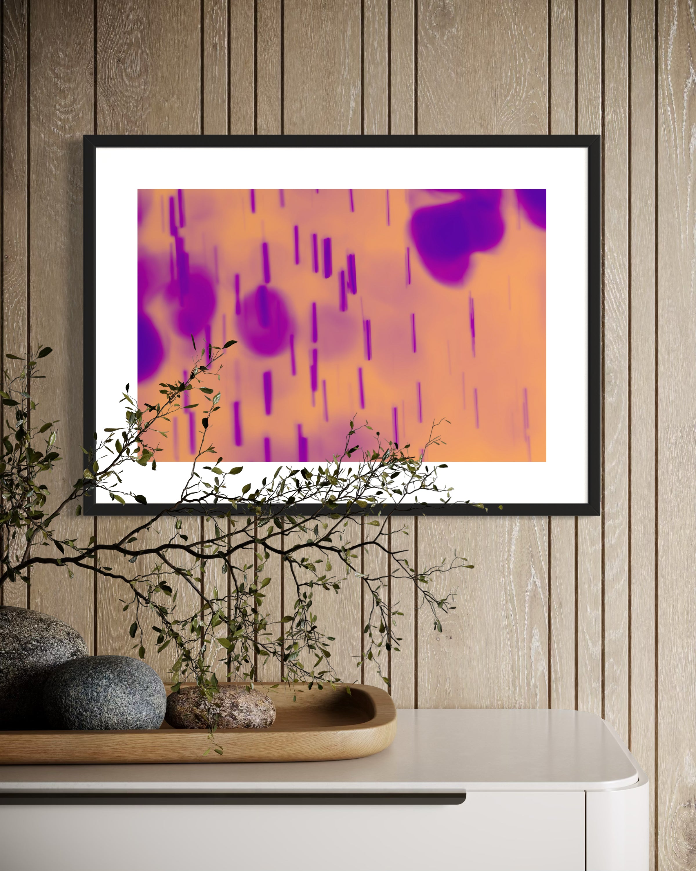 blurred rain in purple on pale yellow background in black frame with white matte on tan wall above white piece of furniture with large twig on top covering part of the framed photo