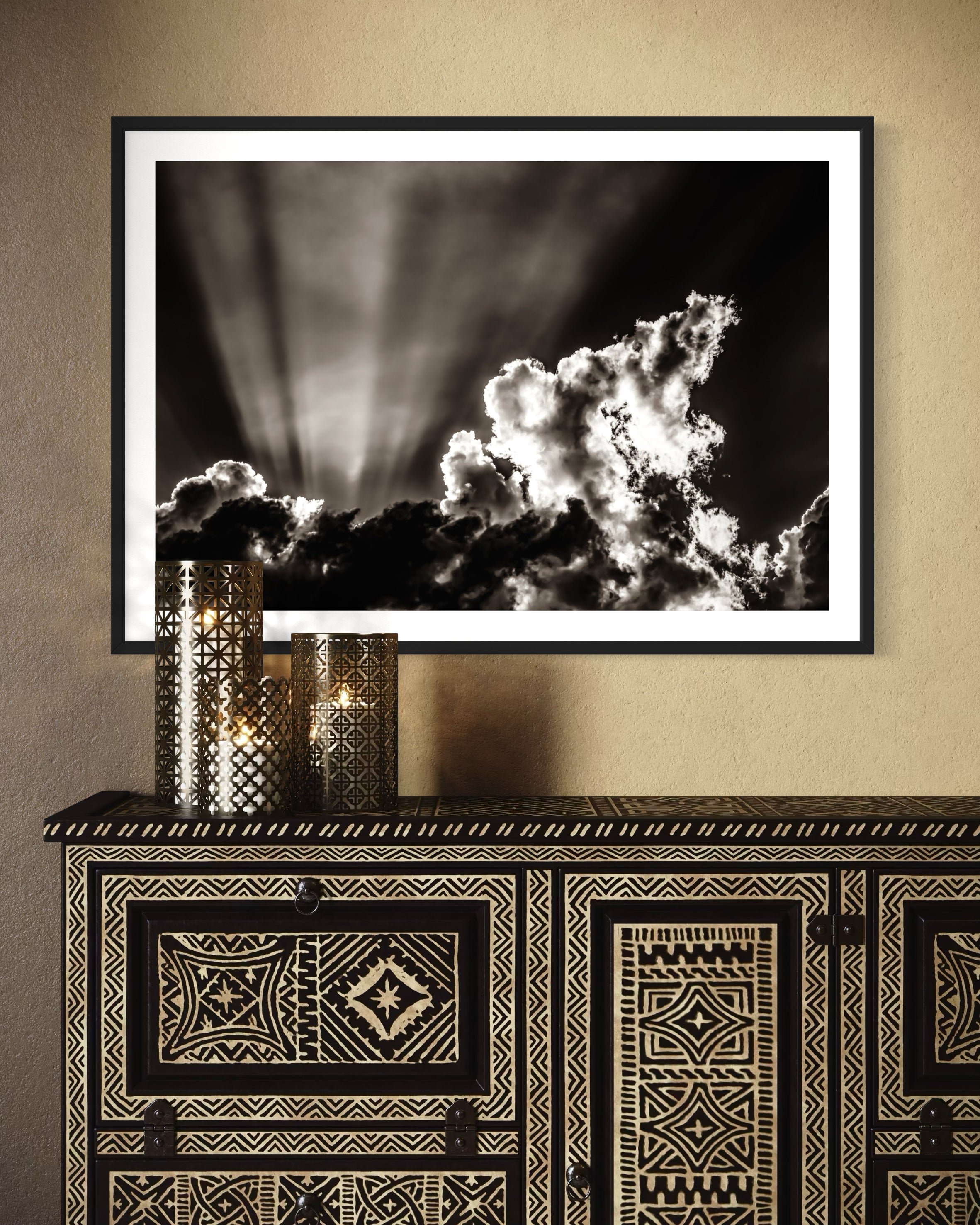 black and white photo of dramatic sky with some clouds and rays of light  in black frame with white matte on yellowish tan wall above southwestern style piece of furniture 