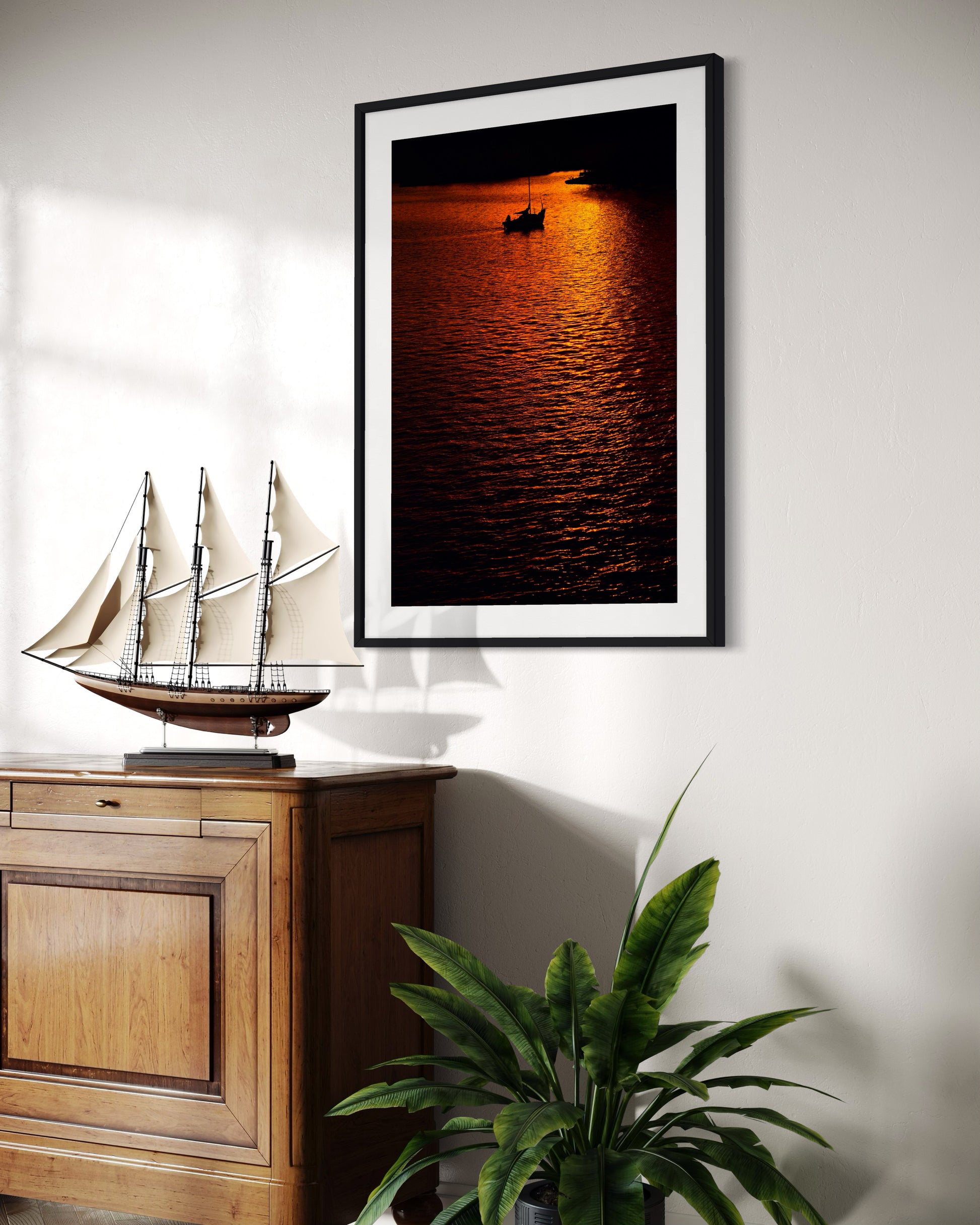 photo of sailboat sailing at sunset, in a very warm tone in black frame with white matte on white wall above small piece of furniture with old ship model on top